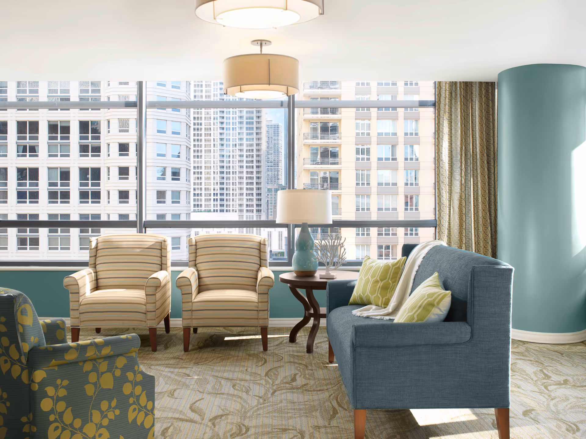 A bright and modern living room area with large windows showing a cityscape view. The room features two striped armchairs, a blue sofa with green and white pillows, a round wooden side table with a lamp and decorative items, and a patterned carpet. The walls are painted in a soft teal color with matching curtains.