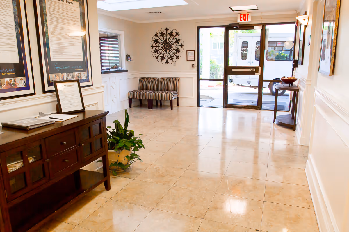 Bright senior living facility lobby with a tiled floor, striped bench, wooden console table, potted plant, wall art, and glass entry doors.