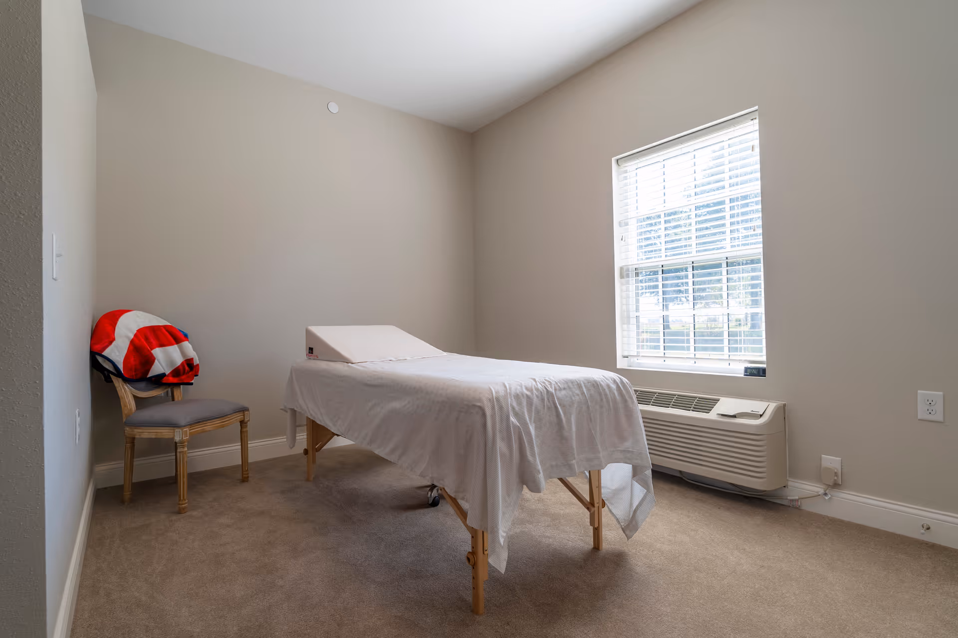 A small room with beige walls and carpeted floor featuring a massage or treatment table covered with a white sheet and a wedge pillow. There is a wooden chair with a red and white blanket draped over it in the corner, and a window with blinds letting in natural light. Below the window is a wall-mounted air conditioning unit.