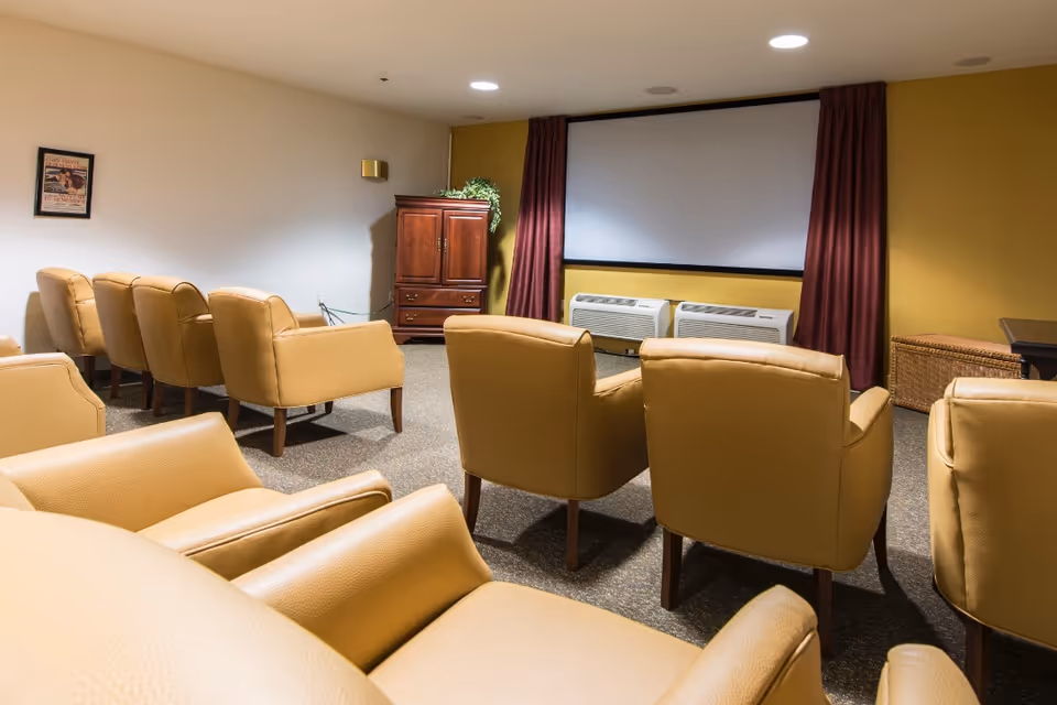 Small screening room with rows of tan armchairs facing a projection screen and wall air conditioners.