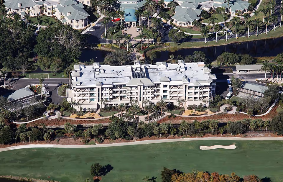 Aerial view of Chateau At Moorings Park senior living facility showing a large multi-story building surrounded by landscaped gardens, palm trees, parking areas, and nearby smaller buildings. A golf course is visible in the foreground.