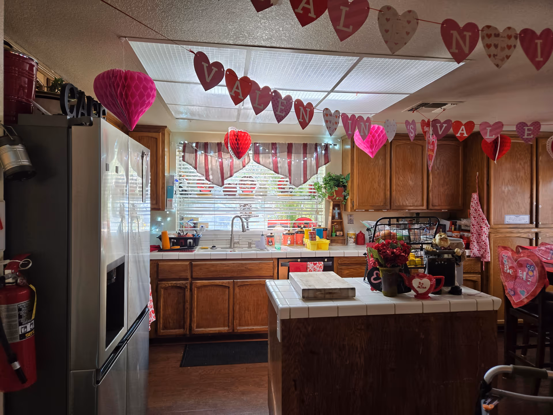 A kitchen decorated with Valentine's Day themed decorations including hanging heart-shaped garlands and paper hearts. The kitchen features wooden cabinets, a white tiled countertop island with a flower pot and other items, a stainless steel refrigerator, a window with striped curtains, and various kitchen utensils and appliances.