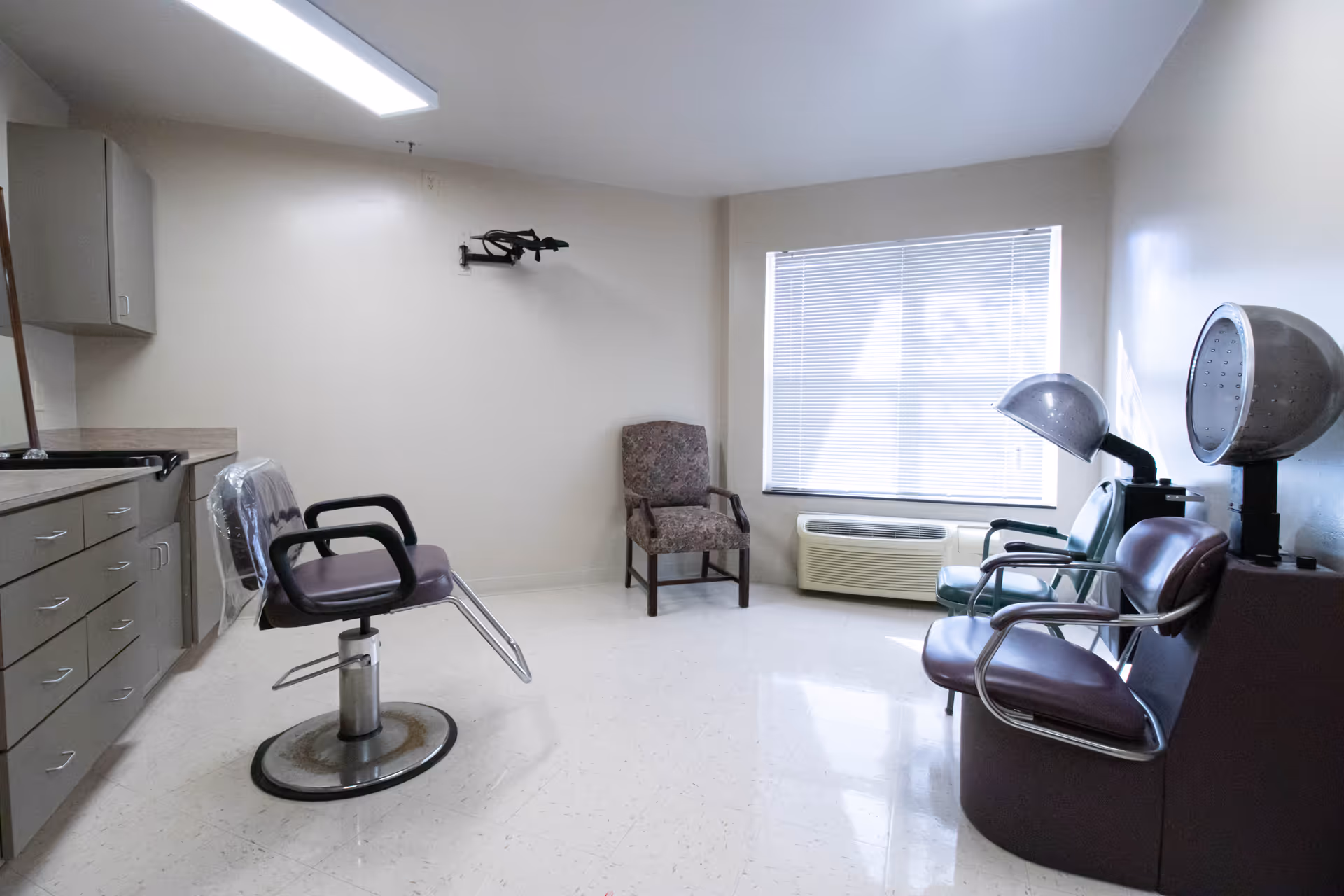 A small salon room with a styling chair in the center, a patterned armchair against the wall, and two vintage hair dryers with attached chairs near a window with closed blinds. There are cabinets and a countertop along the left wall.