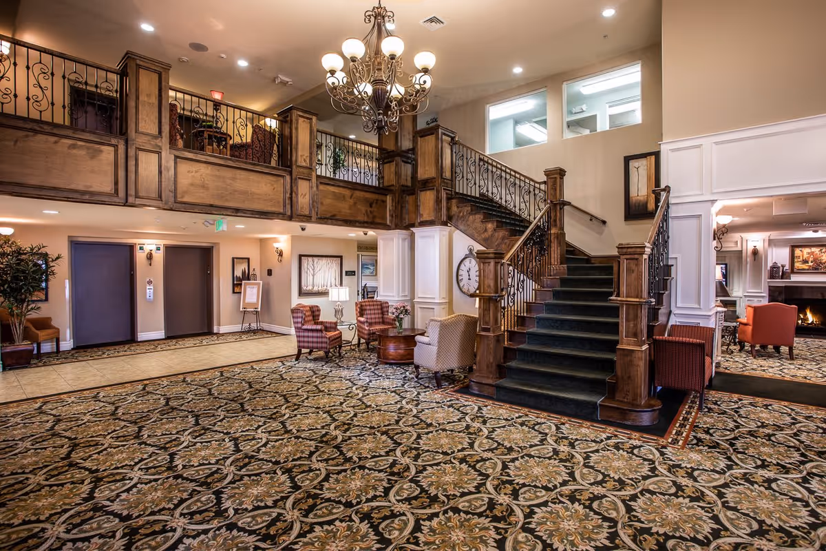 Spacious and elegant senior living facility lobby with a patterned carpet, wooden staircase with ornate railing, chandelier lighting, and comfortable seating areas including armchairs and small tables. There are elevators on the left side and a cozy fireplace area visible on the right.