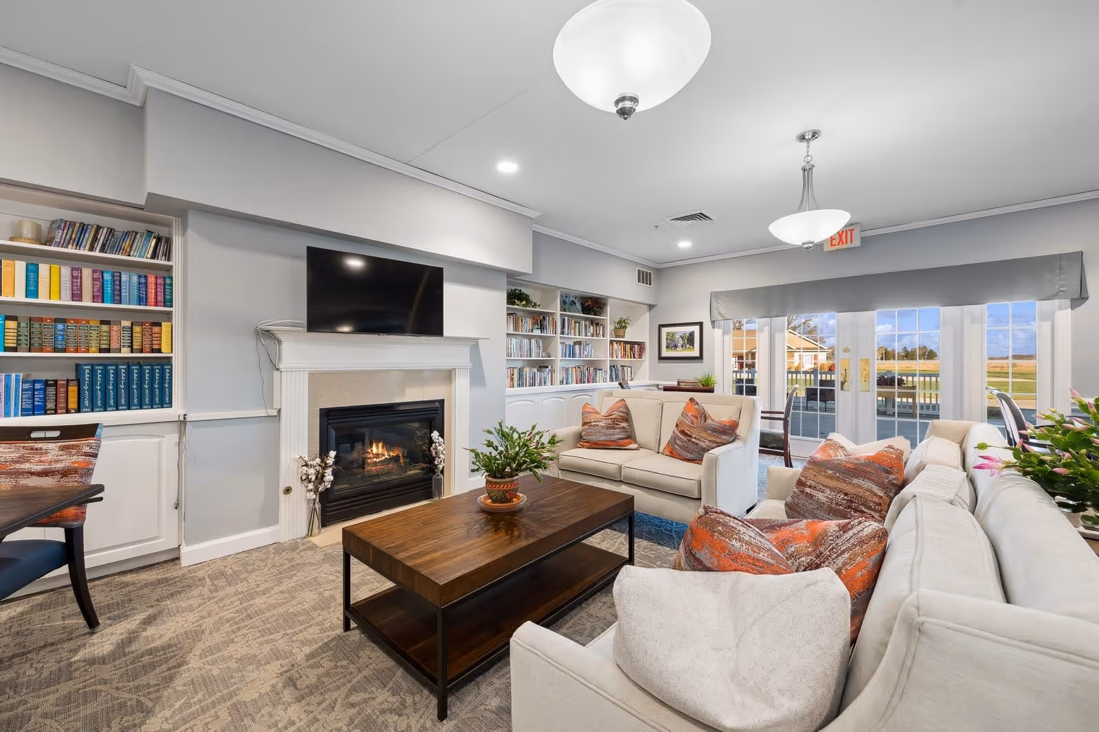 A cozy living room area in Smith Farms Manor featuring two beige sofas with orange and brown patterned pillows arranged around a wooden coffee table. There is a fireplace with a TV mounted above it, built-in bookshelves filled with books, and large glass doors leading to an outdoor patio. The room is well-lit with ceiling lights and has a neutral color scheme with light gray walls and carpeted flooring.