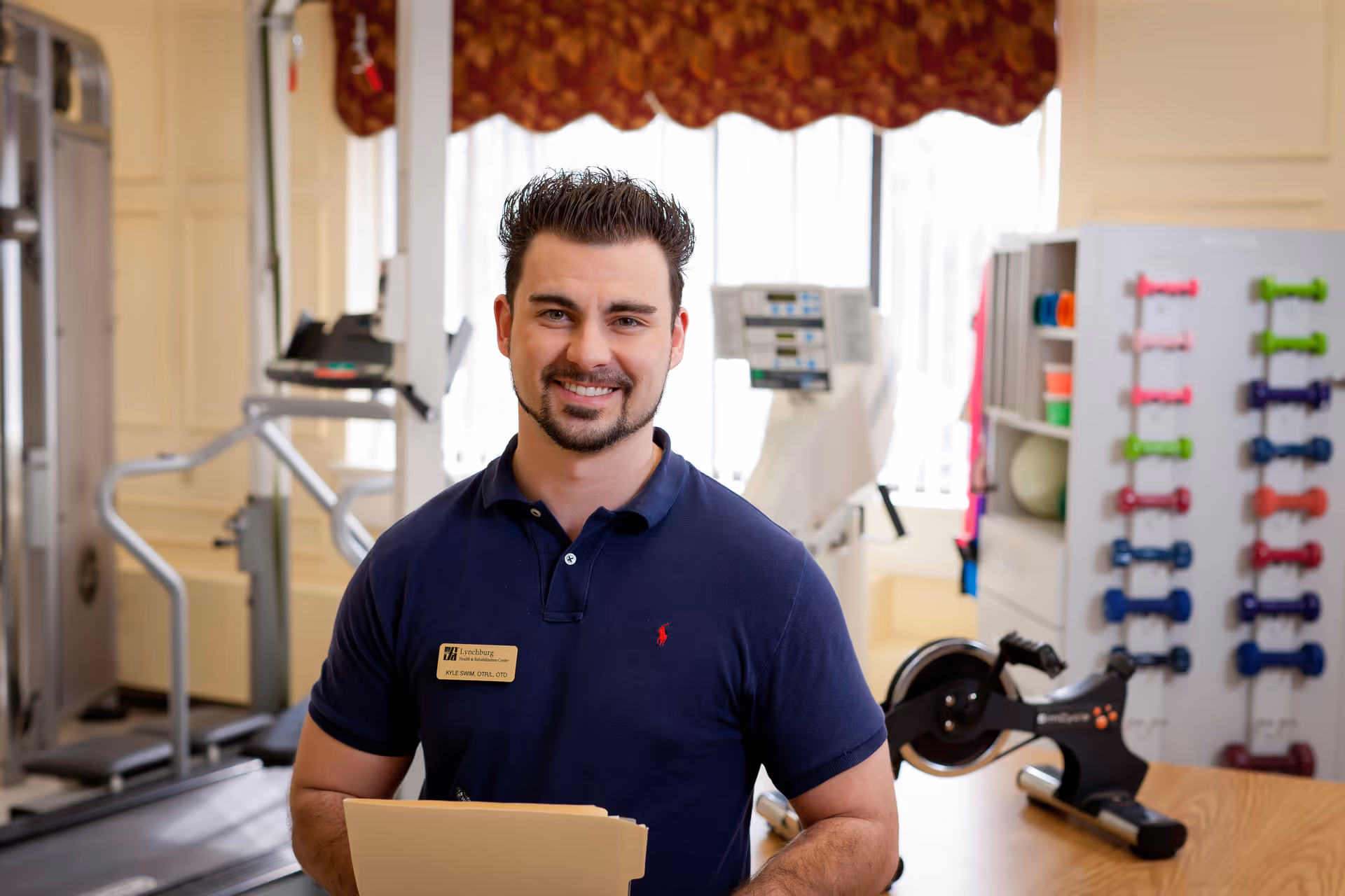 A man wearing a navy blue polo shirt with a name tag stands smiling in a fitness or rehabilitation room. Behind him are exercise equipment including a treadmill, a rowing machine, and a rack of colorful dumbbells. The room is well-lit with large windows and light-colored walls.