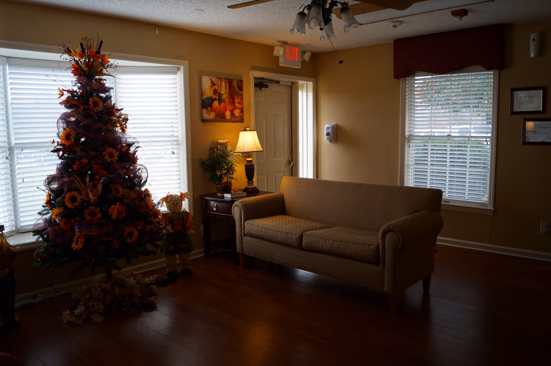 A cozy living room area with a beige patterned sofa, a side table with a lamp and a small plant, a decorated tree with orange flowers and ribbons, and two scarecrow dolls. The room has wooden flooring, two windows with white blinds, a door with an exit sign above it, and framed pictures on the walls.