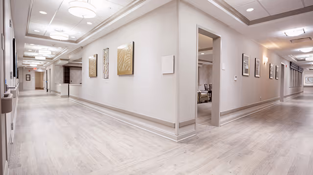 Wide, well-lit interior hallway of a senior living facility with light wood floors, neutral walls, ceiling fixtures, and framed artwork.