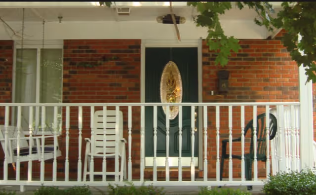 Front porch of a brick building with a white railing, a green door with an oval glass inset, a porch swing and chairs.
