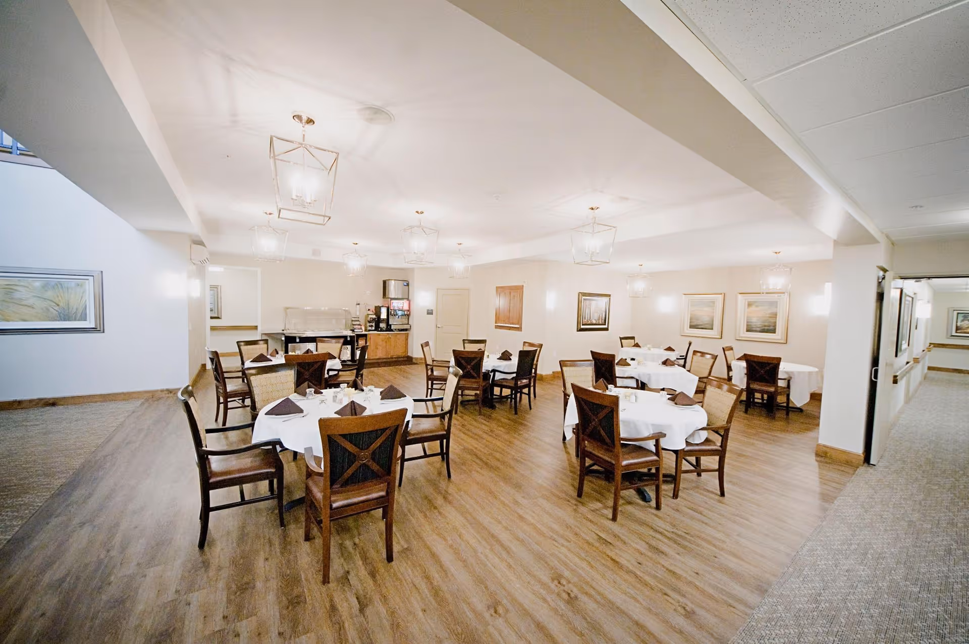 A bright dining room with several round tables covered with white tablecloths and set with brown napkins. Wooden chairs with armrests surround the tables. The room has wood flooring, modern ceiling lights, and framed artwork on the walls. A beverage station is visible in the back corner.