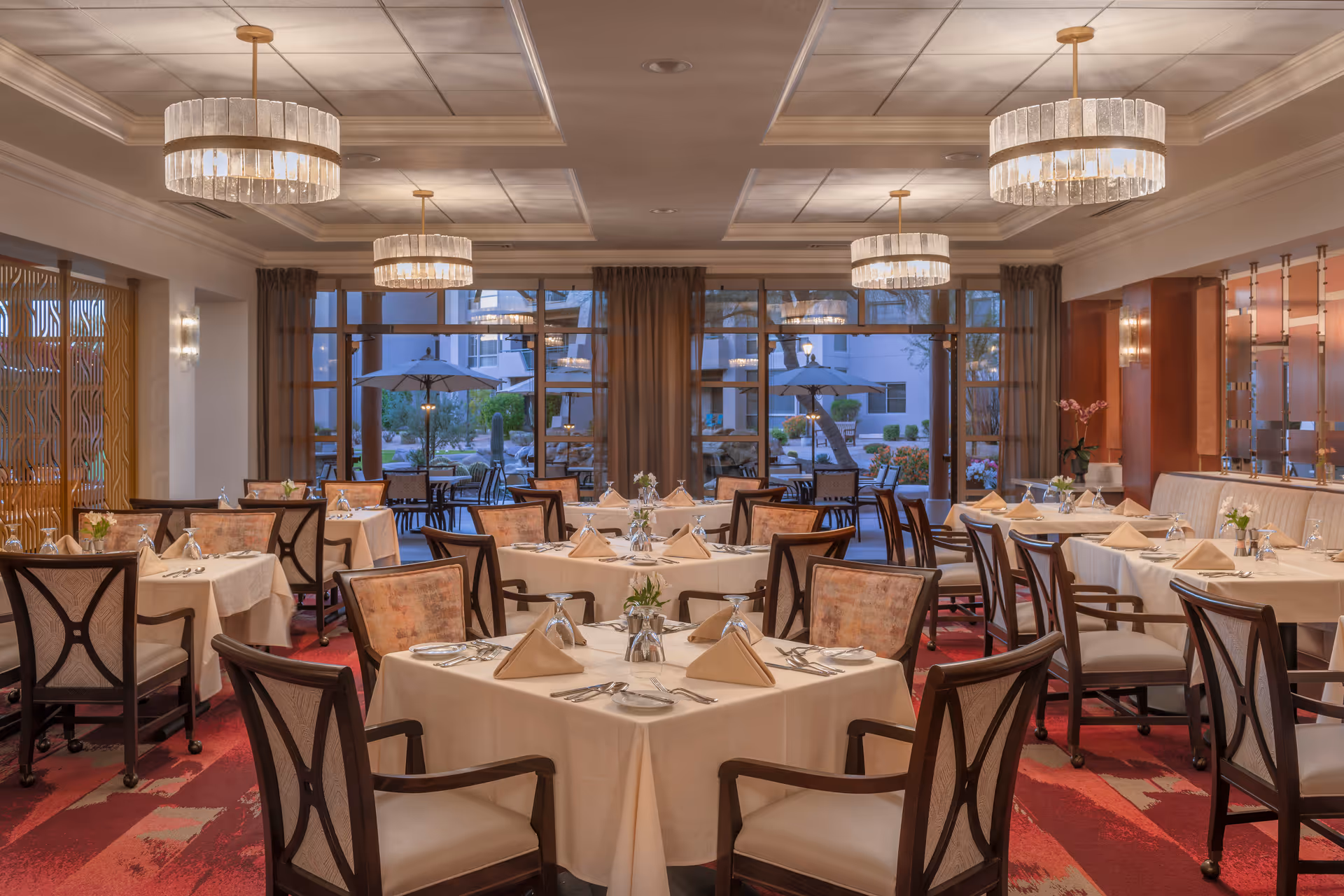 Elegant dining room with multiple tables covered in beige tablecloths, each set with folded napkins, silverware, and small floral centerpieces. The room features large windows with sheer curtains, allowing a view of an outdoor patio area with umbrellas and seating. The ceiling has recessed lighting and decorative chandeliers, and the floor is covered with a red patterned carpet.