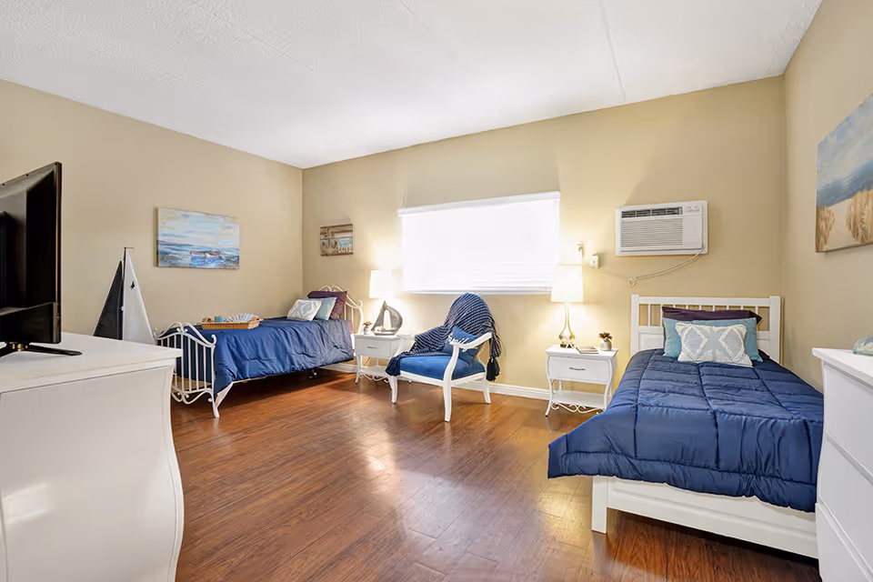 A bright bedroom with two single beds, each with navy blue bedding and decorative pillows. Between the beds is a white nightstand with a lamp and small decor. A blue armchair with a throw blanket is placed near a window with blinds. The room has wooden flooring, beige walls, a wall-mounted air conditioner, and framed artwork on the walls. A white dresser with a TV on top is visible in the foreground.