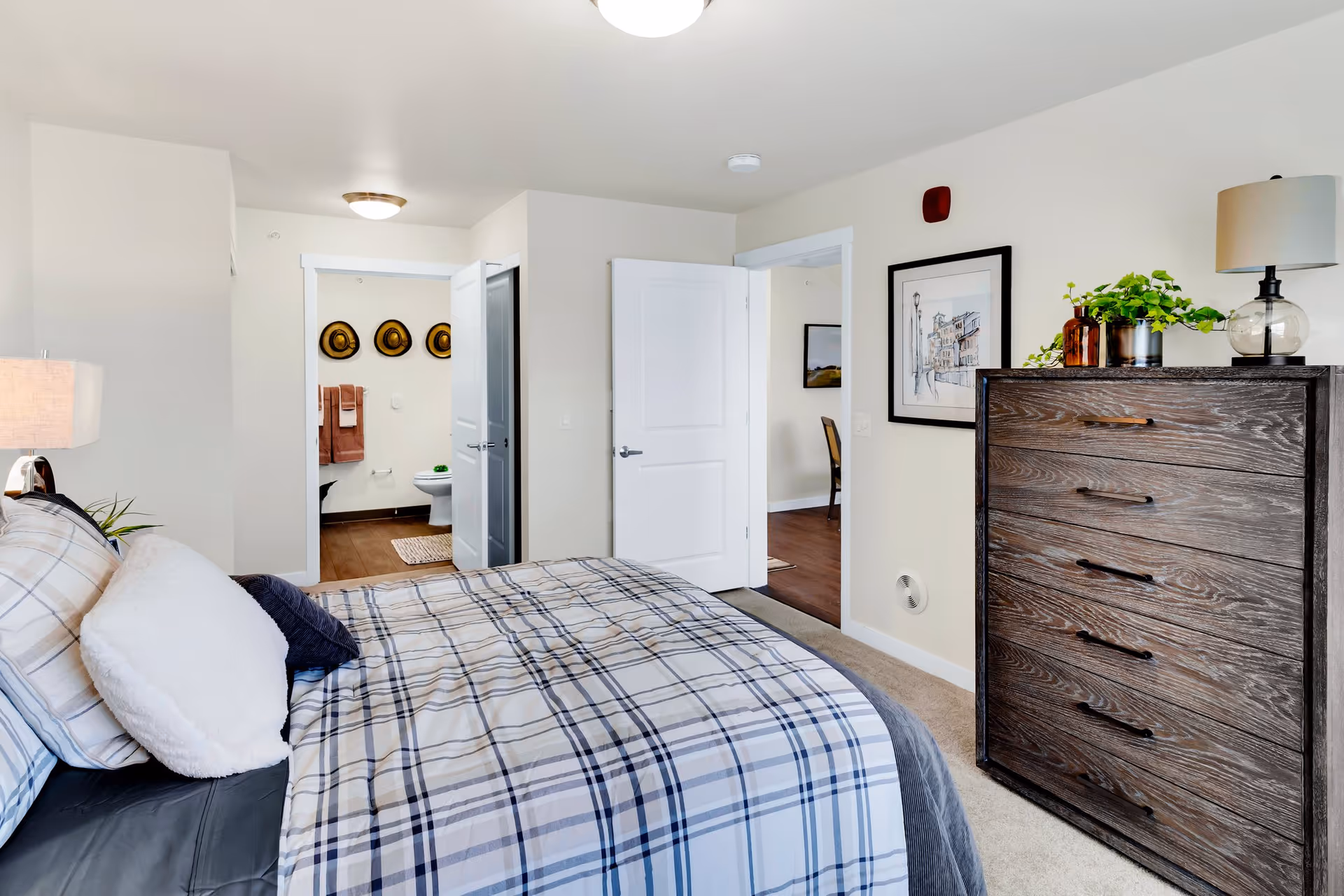 A bedroom with a bed covered in plaid bedding and multiple pillows. To the right is a wooden dresser with a lamp and plants on top. The room has light-colored walls and carpeted floor. Two open doors reveal a bathroom with three hats hanging on the wall and a toilet, and another room with a chair and framed artwork.