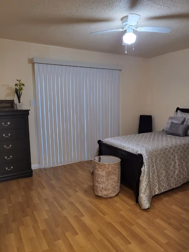 A bedroom with a bed covered in a patterned beige bedspread and multiple pillows. There is a black dresser with drawers on the left side, decorated with a small plant and a framed sign. Vertical blinds cover a window or sliding door behind the bed. A ceiling fan with a light is on the ceiling, and a beige laundry basket with the words 'Home Sweet Home' is placed near the foot of the bed. The floor is wooden.