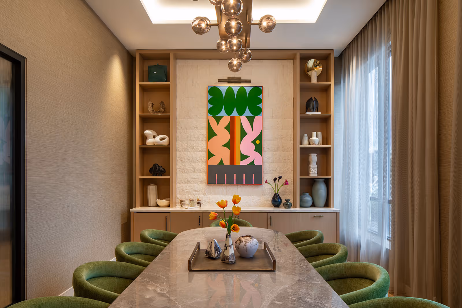 A modern dining room with a long marble table surrounded by green upholstered chairs. The back wall features built-in wooden shelves with decorative items and a colorful abstract painting in the center. A contemporary chandelier hangs from the ceiling, and sheer curtains cover the window on the right side.