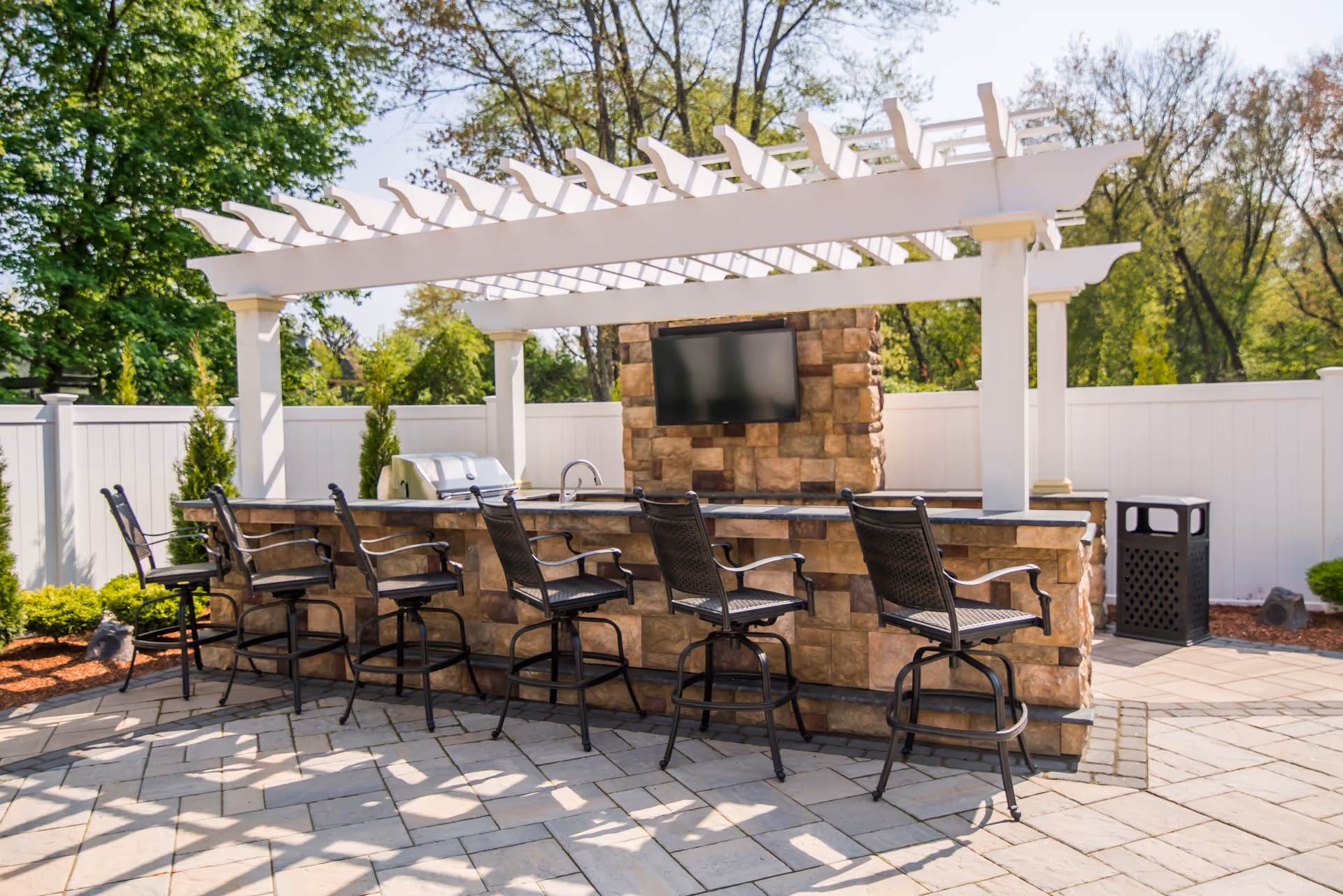 Outdoor patio area with a stone bar counter and six black metal bar stools. Above the bar is a white pergola providing partial shade. A flat-screen TV is mounted on a stone wall behind the bar. There is a grill and a sink built into the bar counter. The area is surrounded by a white privacy fence and greenery.