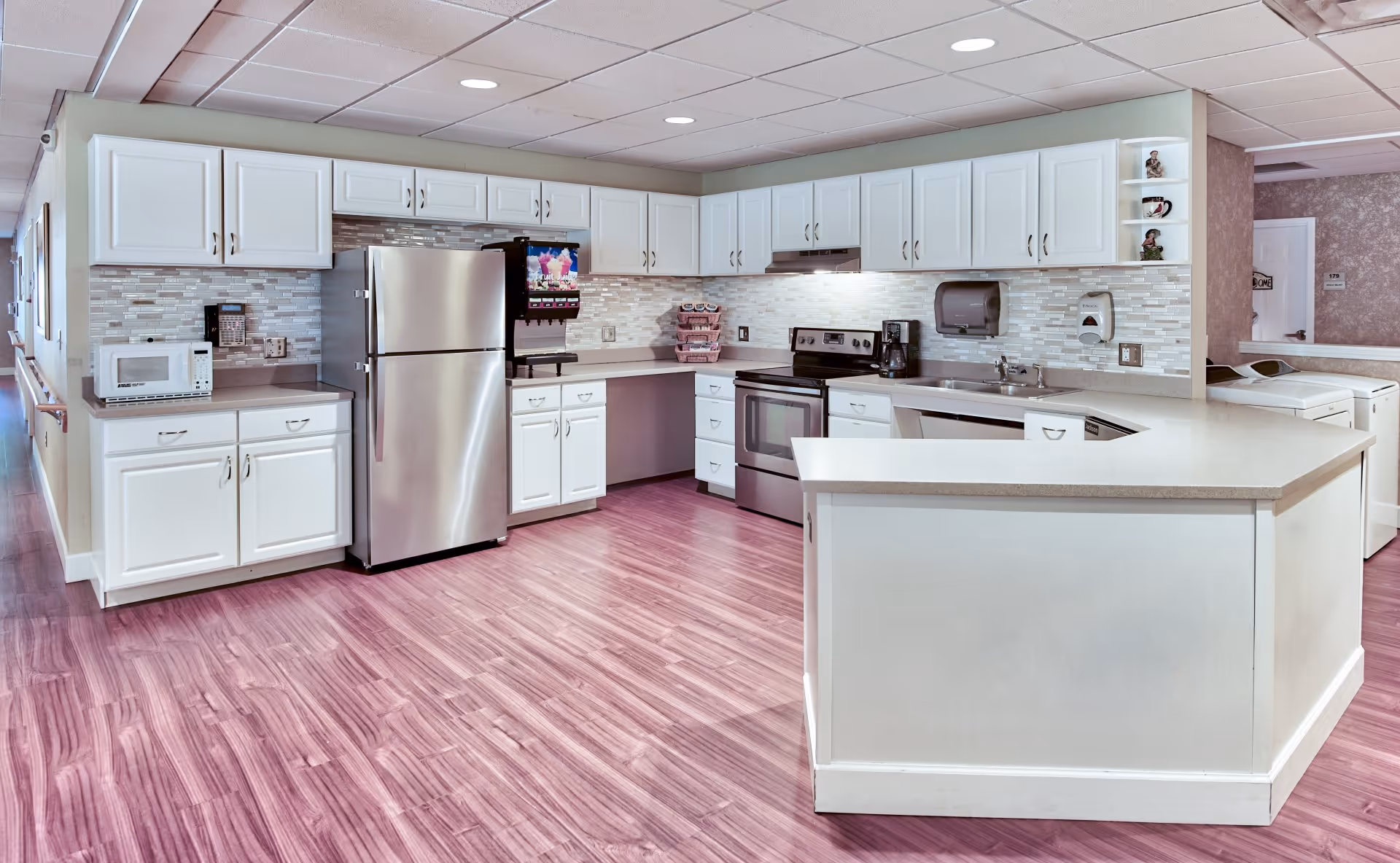 A clean, modern kitchen area in an assisted living facility featuring white cabinets, a stainless steel refrigerator, stove, microwave, coffee maker, and a drink dispenser. The kitchen has a light-colored countertop and a tiled backsplash with a mix of neutral tones. The floor is a polished wood laminate, and the ceiling has recessed lighting. In the background, there is a hallway and a laundry area with washers and dryers.