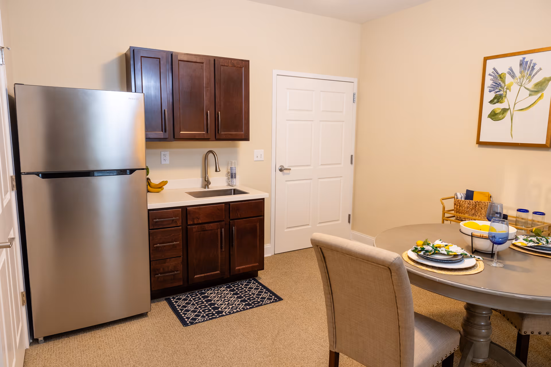 A small kitchen area with dark wooden cabinets, a stainless steel refrigerator, a sink with a modern faucet, and a small blue patterned rug on the floor. Adjacent to the kitchen is a round dining table set with plates, napkins, glasses, and a bowl of lemons. A beige upholstered chair is positioned at the table, and a framed botanical artwork hangs on the wall.