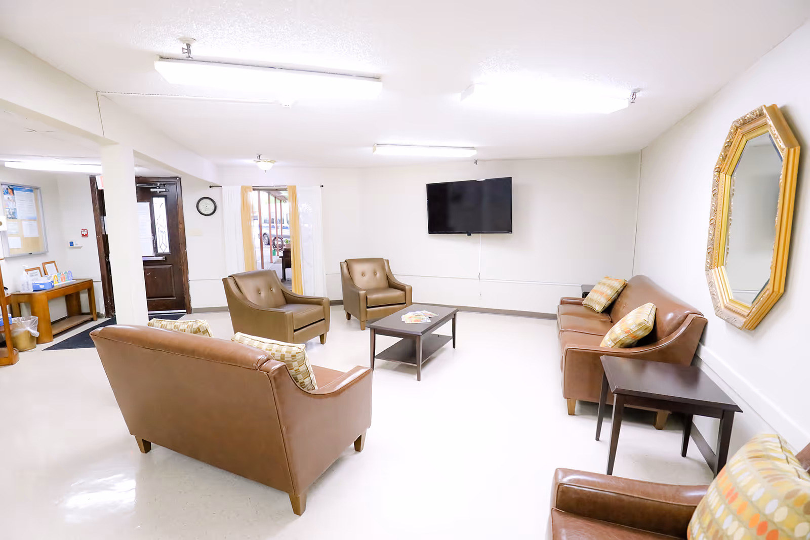 A bright and clean common area with brown leather sofas and armchairs arranged around a small coffee table. A flat-screen TV is mounted on the wall, and a large decorative mirror hangs on another wall. There is a doorway leading outside and a small table with informational materials near the entrance.