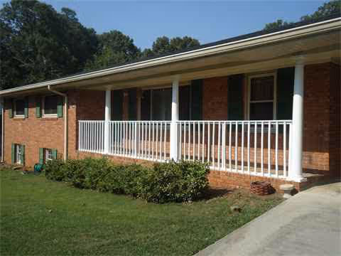 Brick senior living facility exterior with a covered porch, white railings, and surrounding lawn.