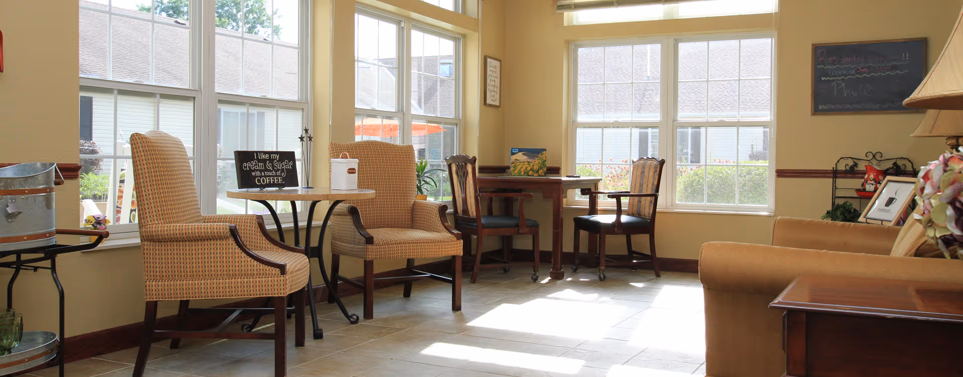 Sunlit common room with upholstered armchairs and small tables arranged by large windows.