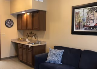 Interior view of a room with a small kitchenette featuring wooden cabinets, a countertop with a sink, and a basket. A dark blue couch with a patterned pillow is positioned next to the kitchenette. A framed artwork hangs on the beige wall above the couch, and a round wall clock is mounted near the kitchenette.