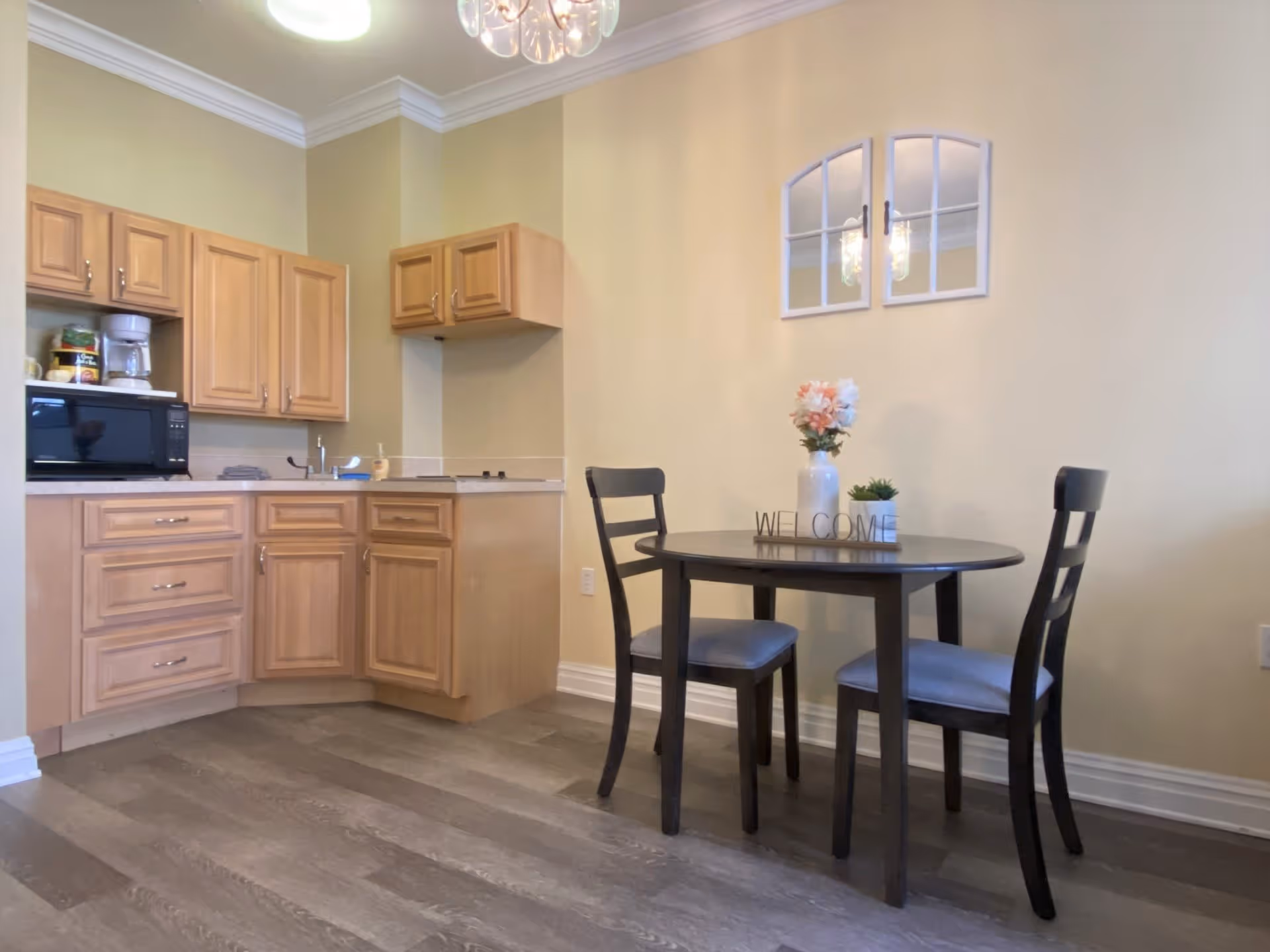 A small kitchen and dining area with light wood cabinets, a microwave, coffee maker, and a small sink. There is a round dark wood table with two matching chairs, a vase with flowers, and a small plant on the table. A decorative wall mirror with an arched windowpane design hangs on the light yellow wall.