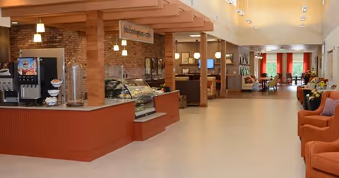 Interior view of a spacious dining area in Stonecroft Health Campus featuring a beverage station with soda dispensers and a display case, wooden pillars, brick walls, and seating areas with orange armchairs and tables in the background.