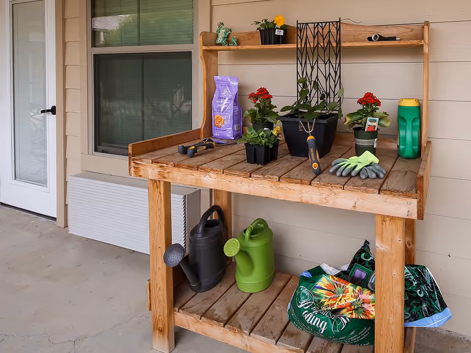 A wooden gardening workbench on a porch with various gardening supplies including potted plants, gardening gloves, small hand tools, watering cans, and bags of soil or fertilizer. The bench is positioned against the exterior wall of a building near a door and window.