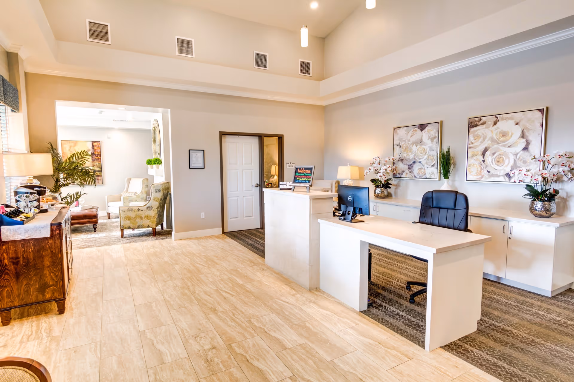 Reception area of The Courte At Citrus Heights featuring a white desk with a computer and office chair, floral artwork on the wall, decorative plants, and a seating area visible through an open doorway.