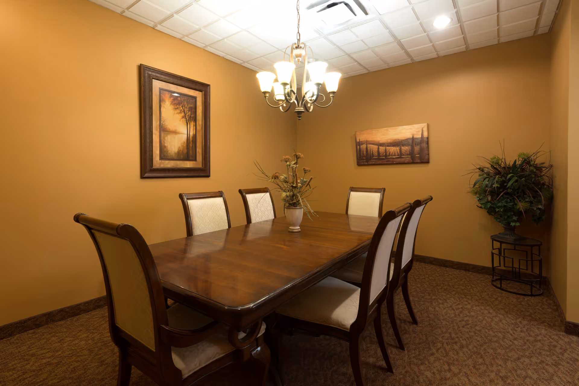 A dining room with a wooden table and six upholstered chairs. The walls are painted a warm beige color and decorated with two framed landscape paintings. A chandelier with multiple lights hangs above the table, and there is a potted plant on a small stand in the corner.