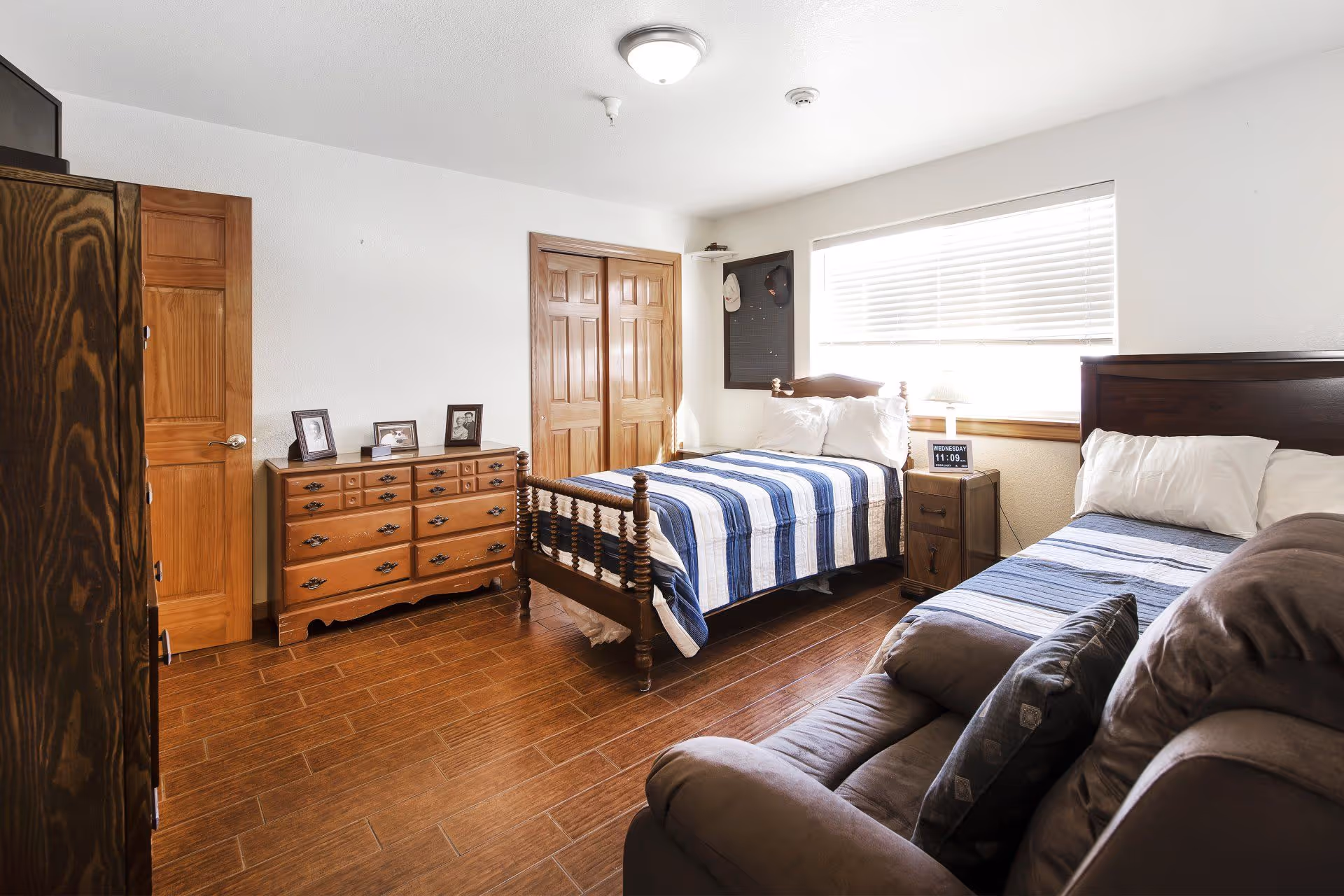 A bright bedroom with two beds, one with a wooden frame and blue-striped bedding, and the other with a dark wooden headboard and similar bedding. There is a wooden dresser with framed photos on top, a wooden wardrobe, a brown recliner chair, and a window with blinds letting in natural light. A small nightstand with a digital clock is between the beds.