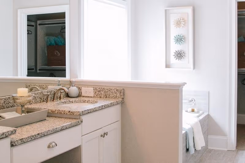 Bright bathroom with a granite countertop sink, a large mirror, and a candle on the counter. In the background, there is a bathtub with a towel hanging over the edge and a small tray with soap. The room has white walls and a window letting in natural light.