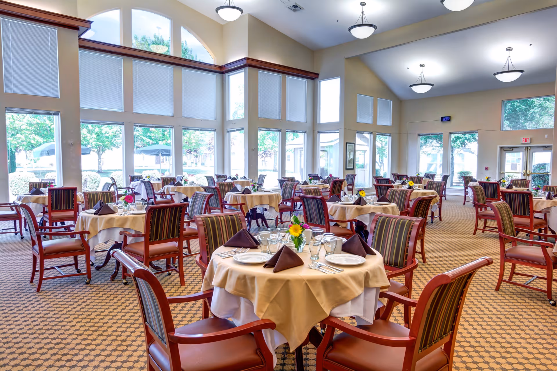 A bright and spacious dining room with multiple round tables covered with beige tablecloths and set with plates, glasses, silverware, and folded brown napkins. Each table has a small flower arrangement. The room features large windows with white blinds, allowing natural light to fill the space. The chairs have wooden frames with striped upholstery. Ceiling lights hang from the high ceiling.