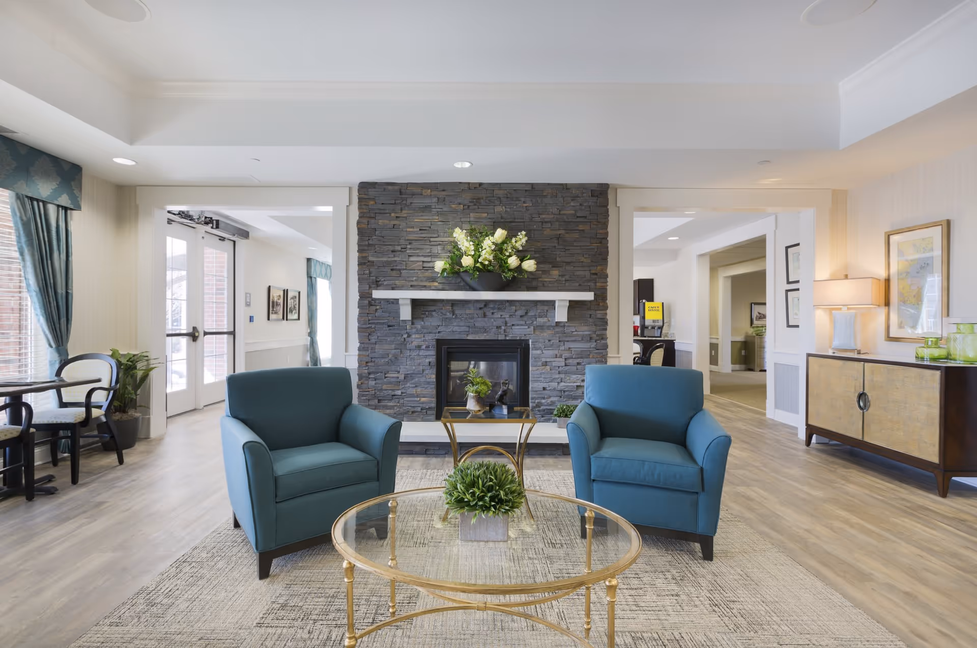 A bright and spacious living room area with two blue armchairs facing a stone fireplace adorned with a flower arrangement. A glass coffee table with a small green plant is placed in front of the chairs on a patterned rug. The room features wood flooring, large windows with blue curtains, and a sideboard with a lamp and decorative items. Doors and hallways lead to other parts of the facility.