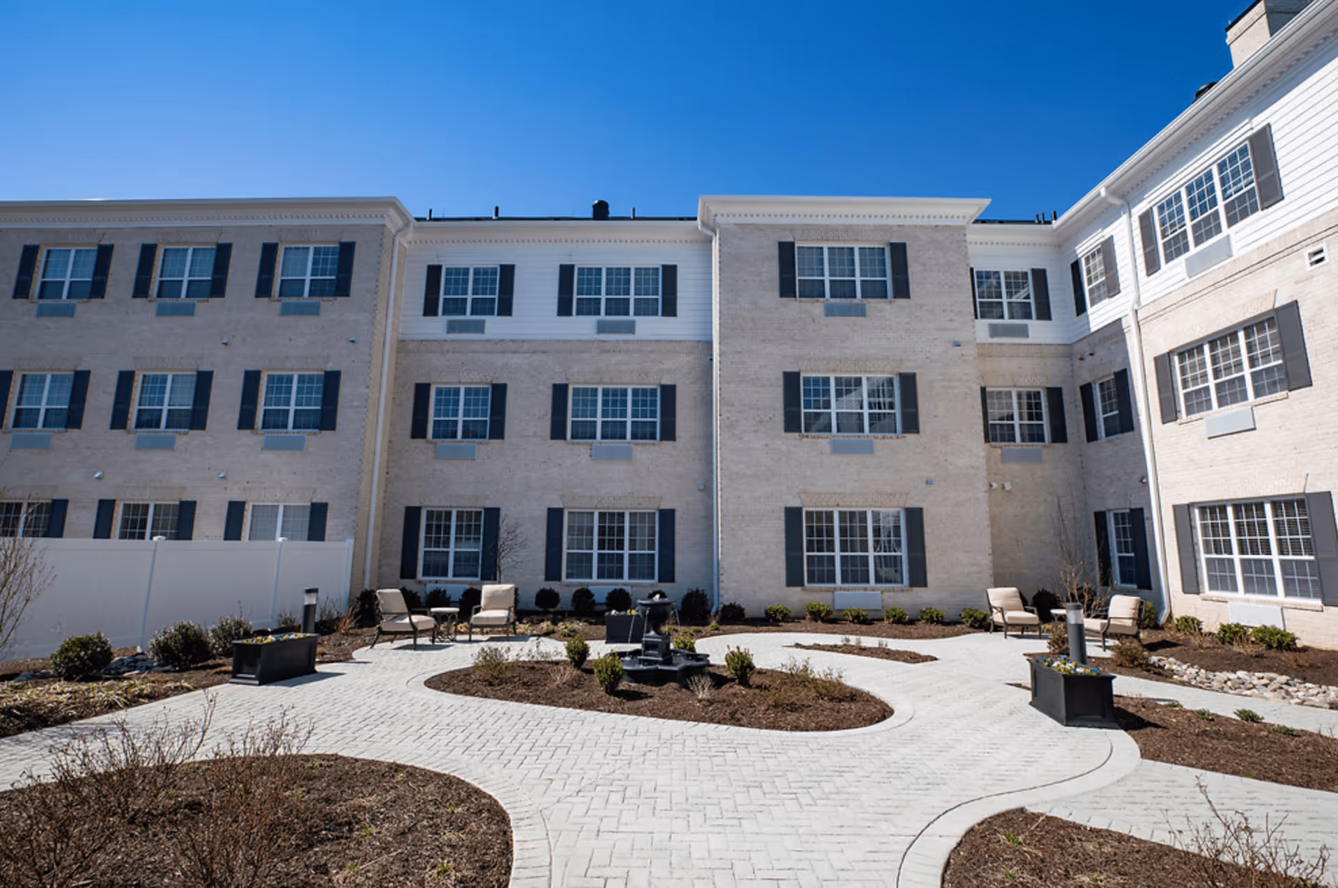 Outdoor courtyard area of a senior living facility with a paved walkway, garden beds, several chairs, and a small water fountain in the center. The building surrounding the courtyard is three stories tall with multiple windows and light-colored brick exterior under a clear blue sky.