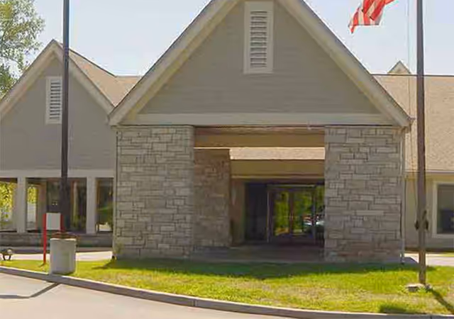 Front exterior view of Sherbrooke Village Living Center showing a stone and beige building entrance with a peaked roof, two flagpoles with an American flag on the right, and a driveway in front.