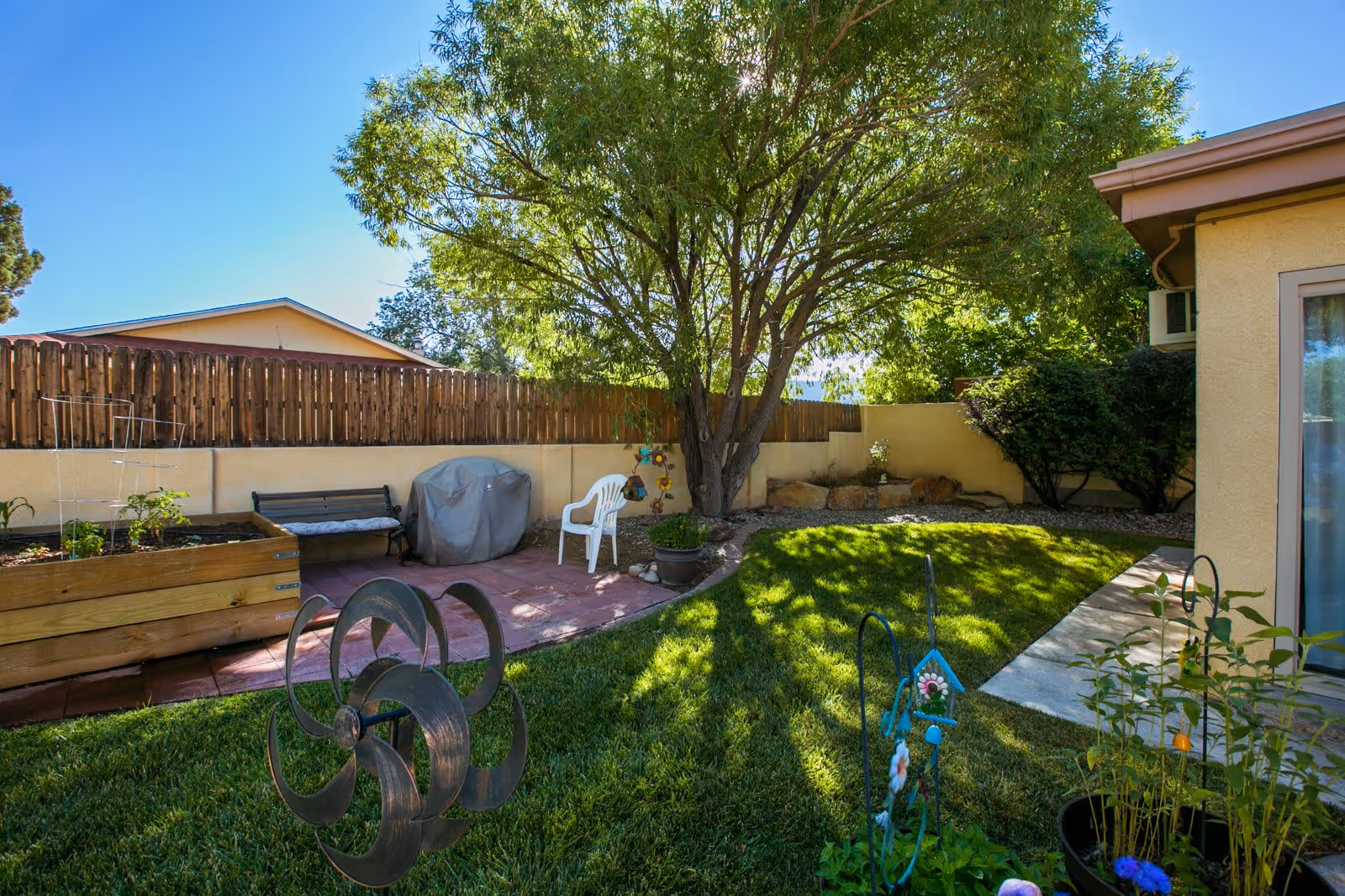 A sunny outdoor garden area with a large tree providing shade, a wooden raised garden bed, a bench, a covered grill, a white plastic chair, and various plants and garden decorations. The area is enclosed by a wooden fence and a beige wall, with a building partially visible on the right side.