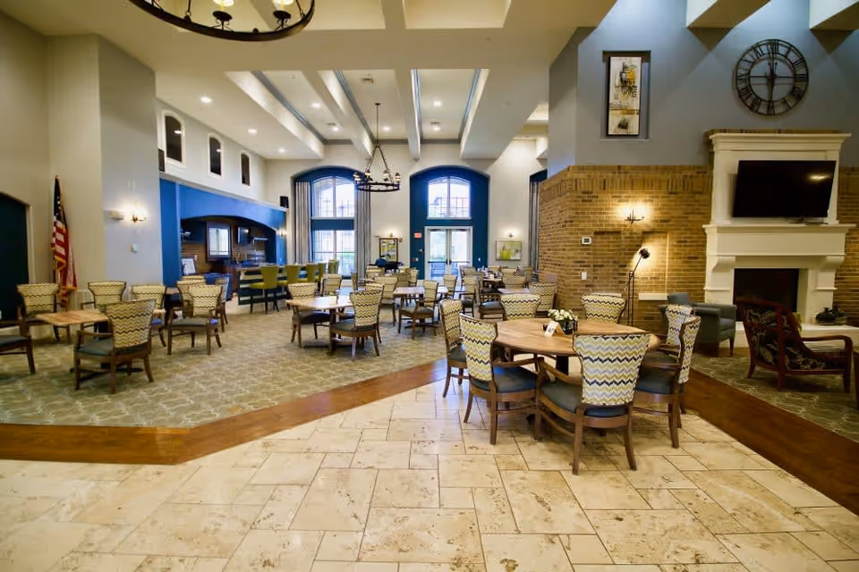 Spacious dining and common area with multiple round tables and cushioned chairs arranged neatly. The room features high ceilings with exposed beams, large arched windows letting in natural light, a fireplace with a mounted TV above it, and a bar area with high stools. The decor includes a mix of carpet and tile flooring, wall art, and a large clock above the fireplace.