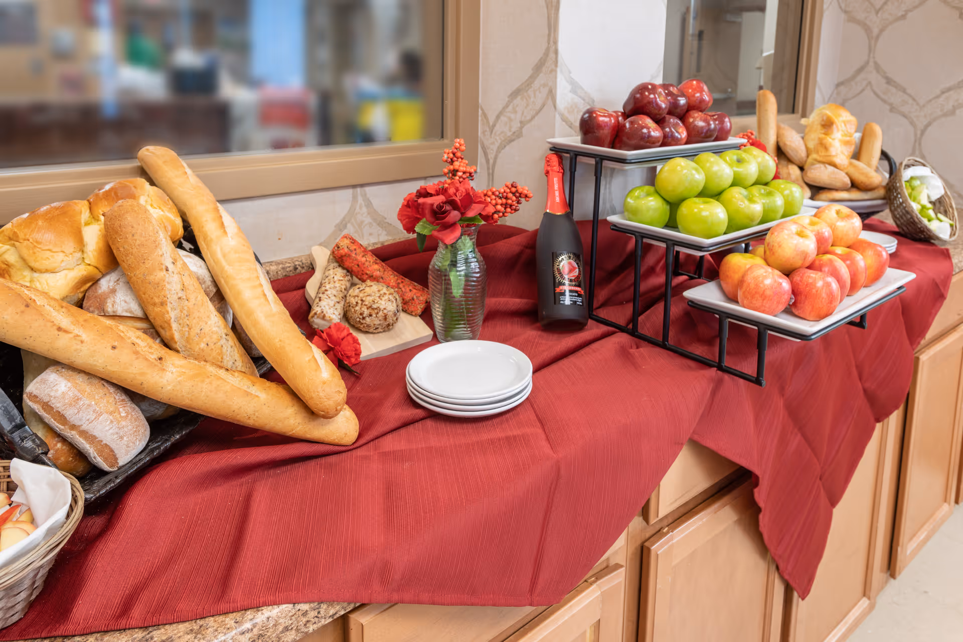 A buffet table covered with a red tablecloth displaying various breads including baguettes and rolls, a tiered stand with red, green, and pink apples, a bottle of sparkling beverage, a small vase with red flowers, and a stack of white plates.