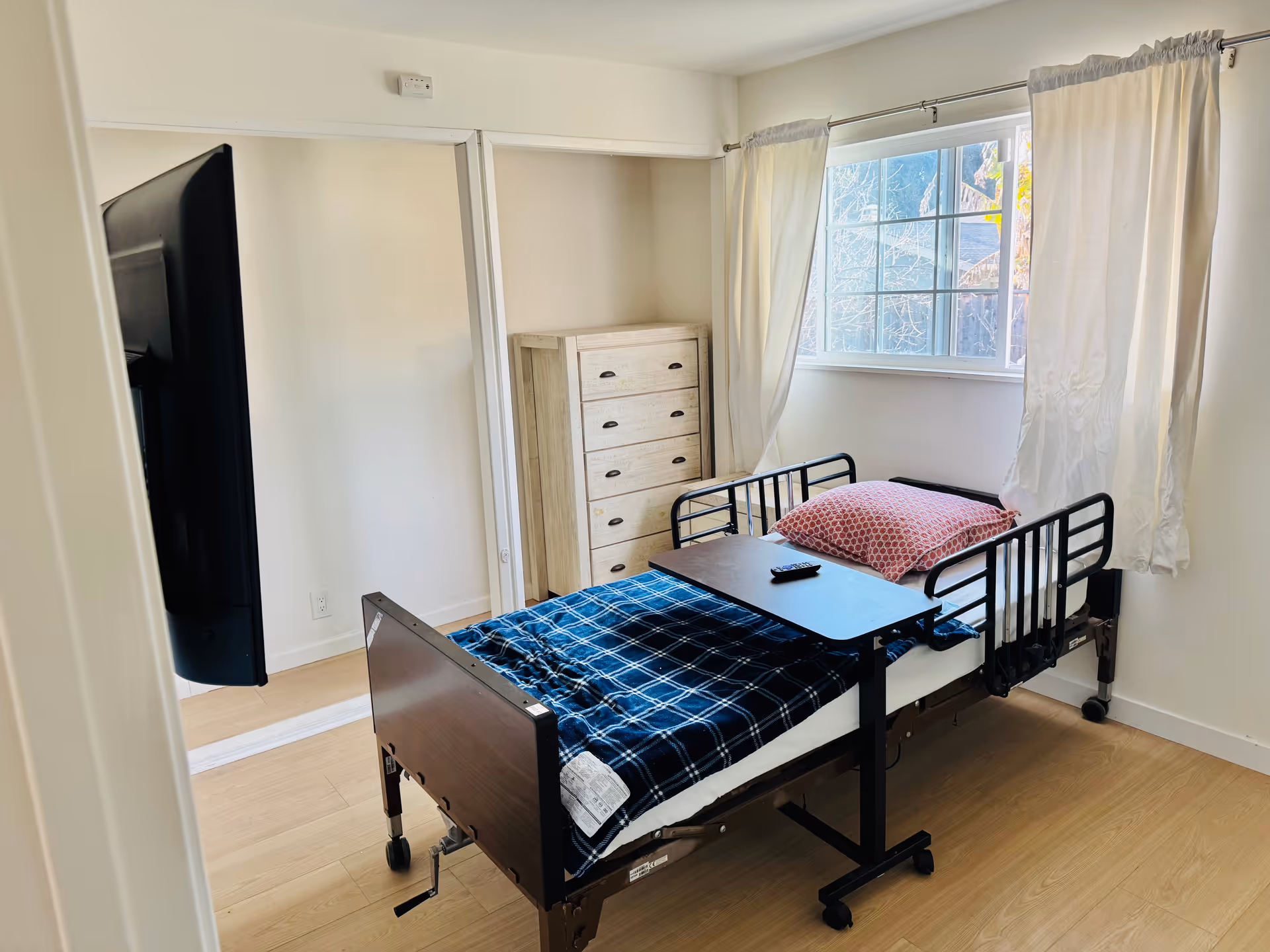 A small bedroom in an elderly care facility featuring a hospital-style bed with side rails, a blue plaid blanket, and a pink patterned pillow. A black overbed table with a remote control rests on the bed. There is a wooden chest of drawers against the wall and a large window with white curtains allowing natural light into the room. A flat-screen TV is mounted on the wall opposite the bed.