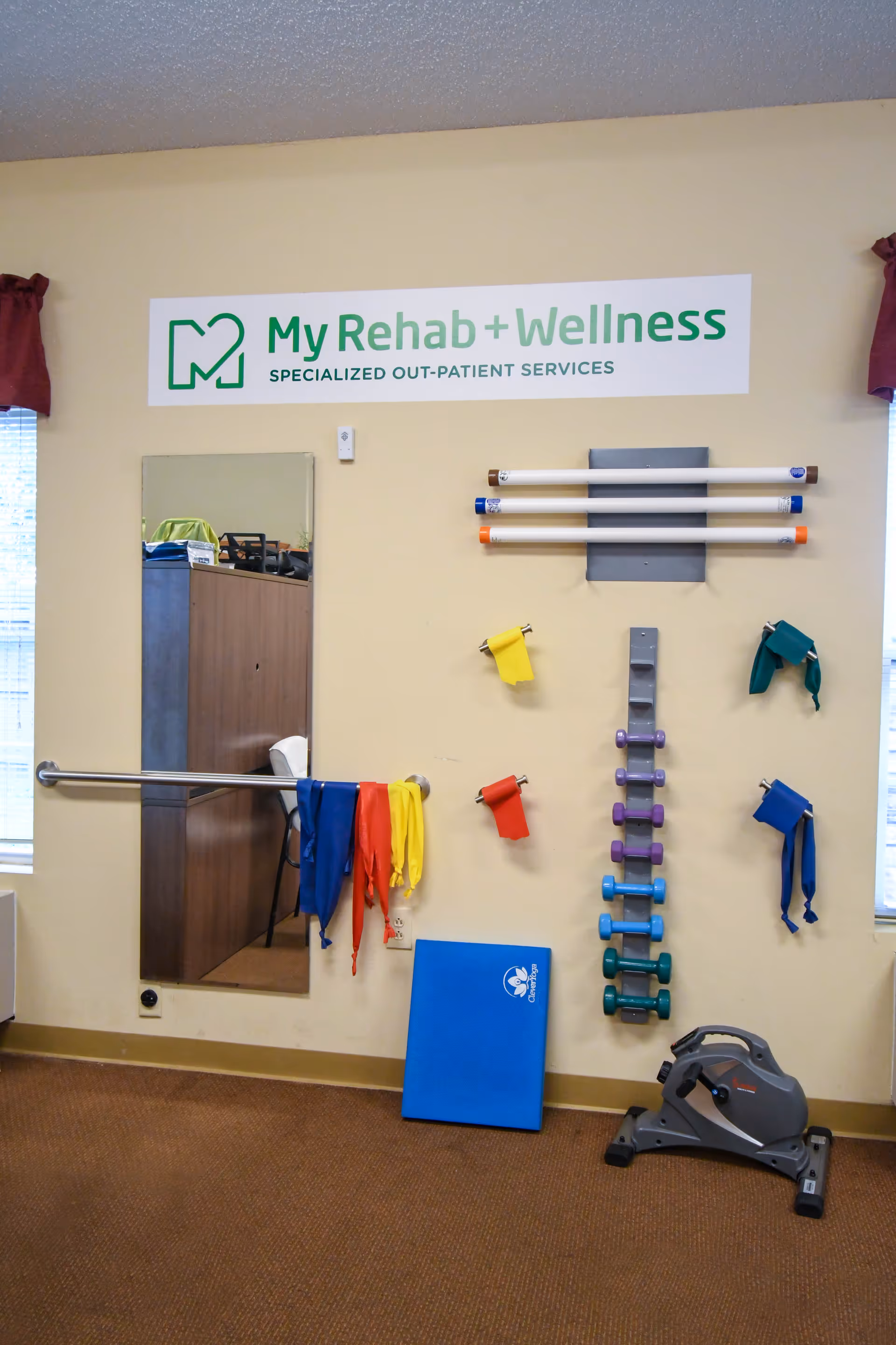 A rehabilitation and wellness room with exercise equipment including resistance bands, dumbbells, a blue exercise mat, and a pedal exerciser. A mirror is mounted on the wall next to a handrail, and a sign above reads 'My Rehab + Wellness Specialized Out-Patient Services'.