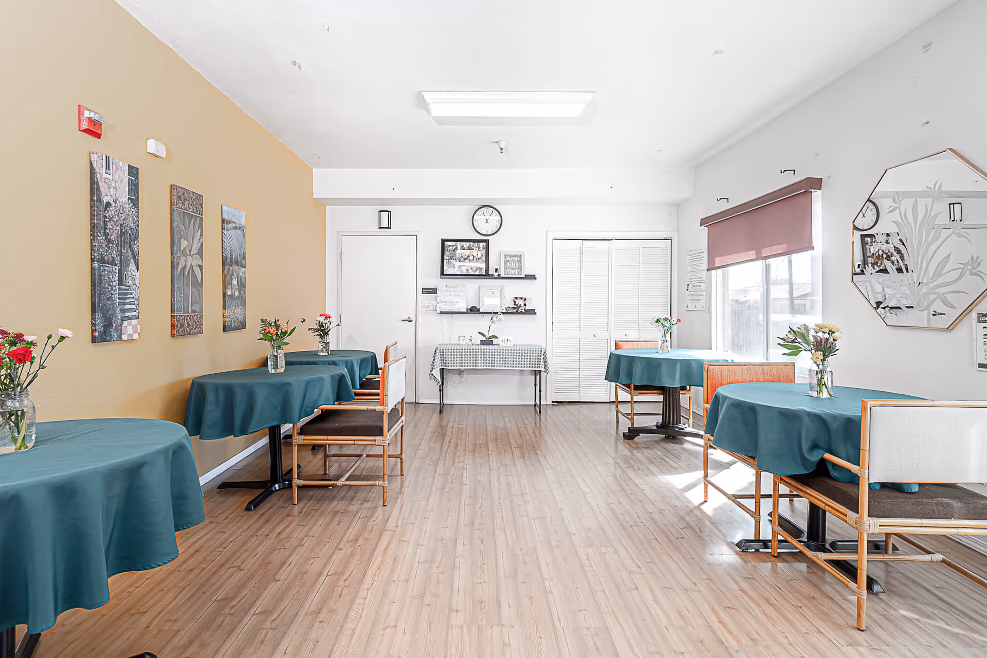 A bright dining room with wooden flooring and several round tables covered with teal tablecloths. Each table has a small vase with flowers. The walls are painted white and light yellow, decorated with framed pictures and a large mirror. There are wooden chairs with cushions around the tables, a clock on the wall, and a window with a purple blind letting in natural light.