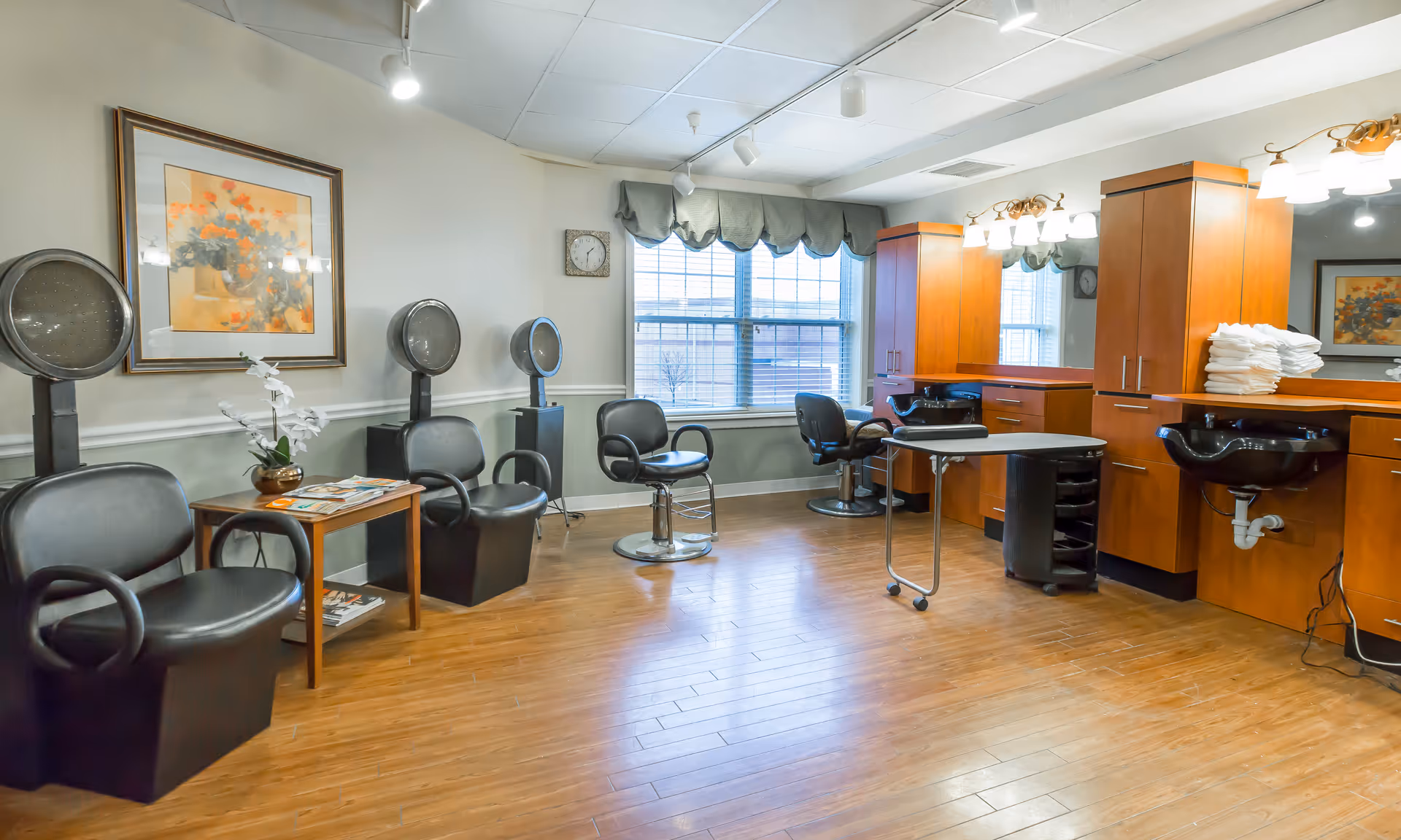 Interior view of a salon area in Pegasus Landing of Overland Park featuring multiple black salon chairs, hair drying stations, a wooden floor, a large window with a valance, wooden cabinets with mirrors and lights, and a small table with magazines and a flower vase.