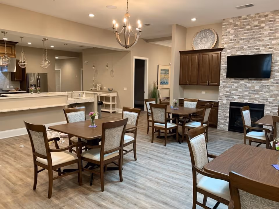 Well-lit communal dining area with multiple wooden tables and chairs, a kitchenette in the background, and a wall-mounted TV above a fireplace.