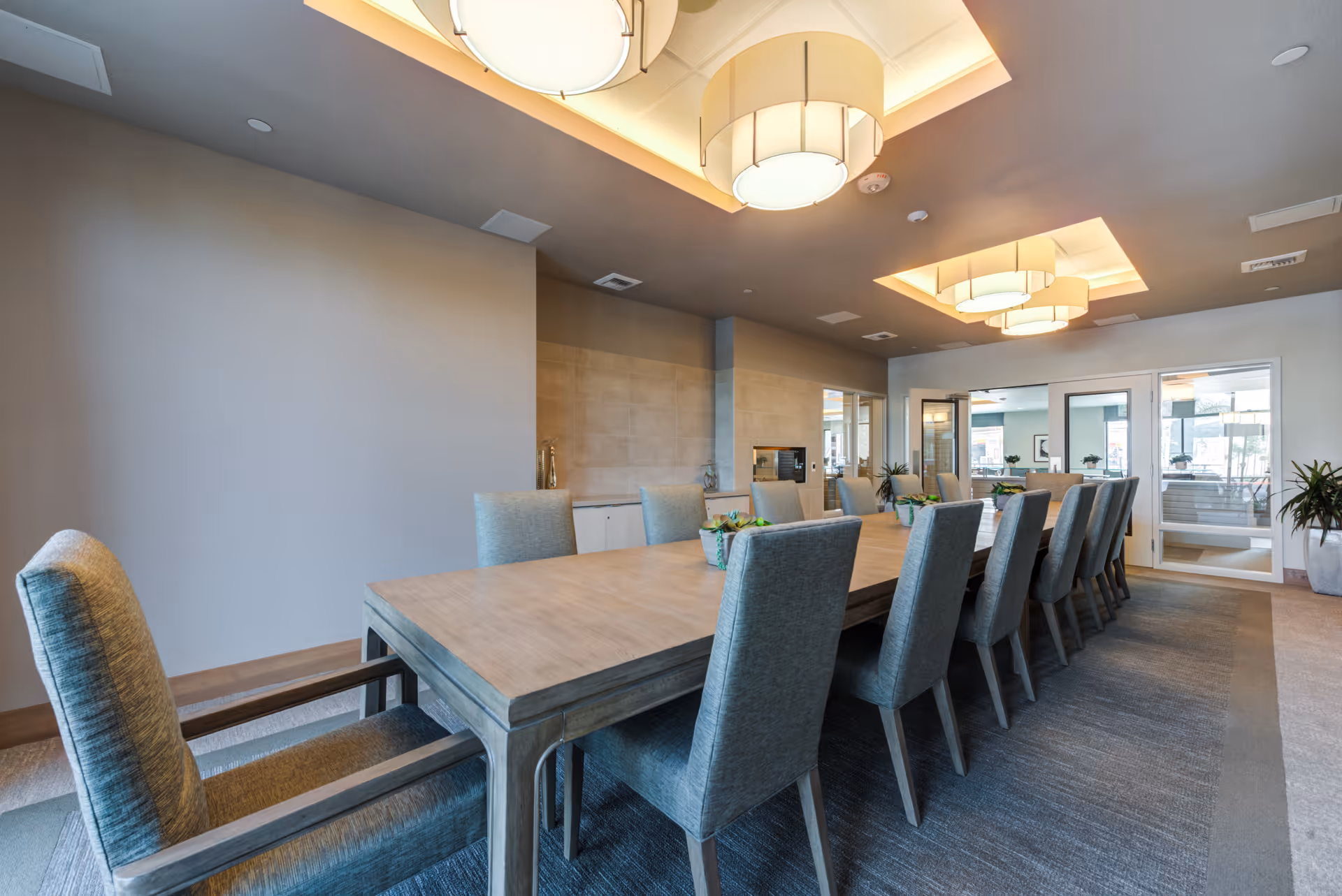 A modern dining room with a long wooden table surrounded by upholstered chairs. The room features large ceiling light fixtures, neutral-colored walls, and a carpeted floor. There are glass doors at the far end leading to another area, and some decorative plants are placed around the room.