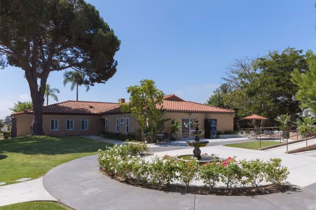 Single-story building with a red tile roof surrounded by green lawns, trees, and a circular flower bed with a small fountain in the center. There is a paved walkway leading to the entrance, and outdoor seating with umbrellas is visible on the right side.
