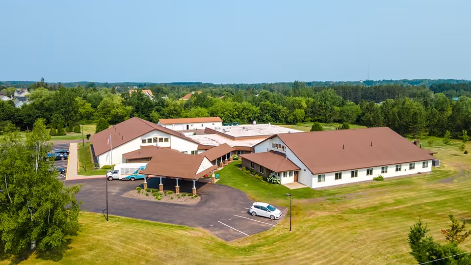 Aerial view of a senior living facility named Gardenview, showing multiple connected buildings with brown roofs surrounded by green lawns and trees. There is a parking area with a few cars and a covered entrance at the front of the building.
