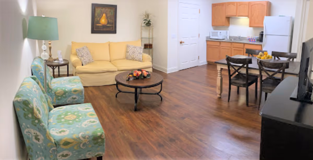 A cozy living area with a yellow sofa, two patterned blue chairs, a round wooden coffee table with a floral centerpiece, and a side table with a lamp. In the background, there is a small kitchen with wooden cabinets, a white refrigerator, microwave, and a dining table with four chairs. The floor is wooden and the walls are painted white.
