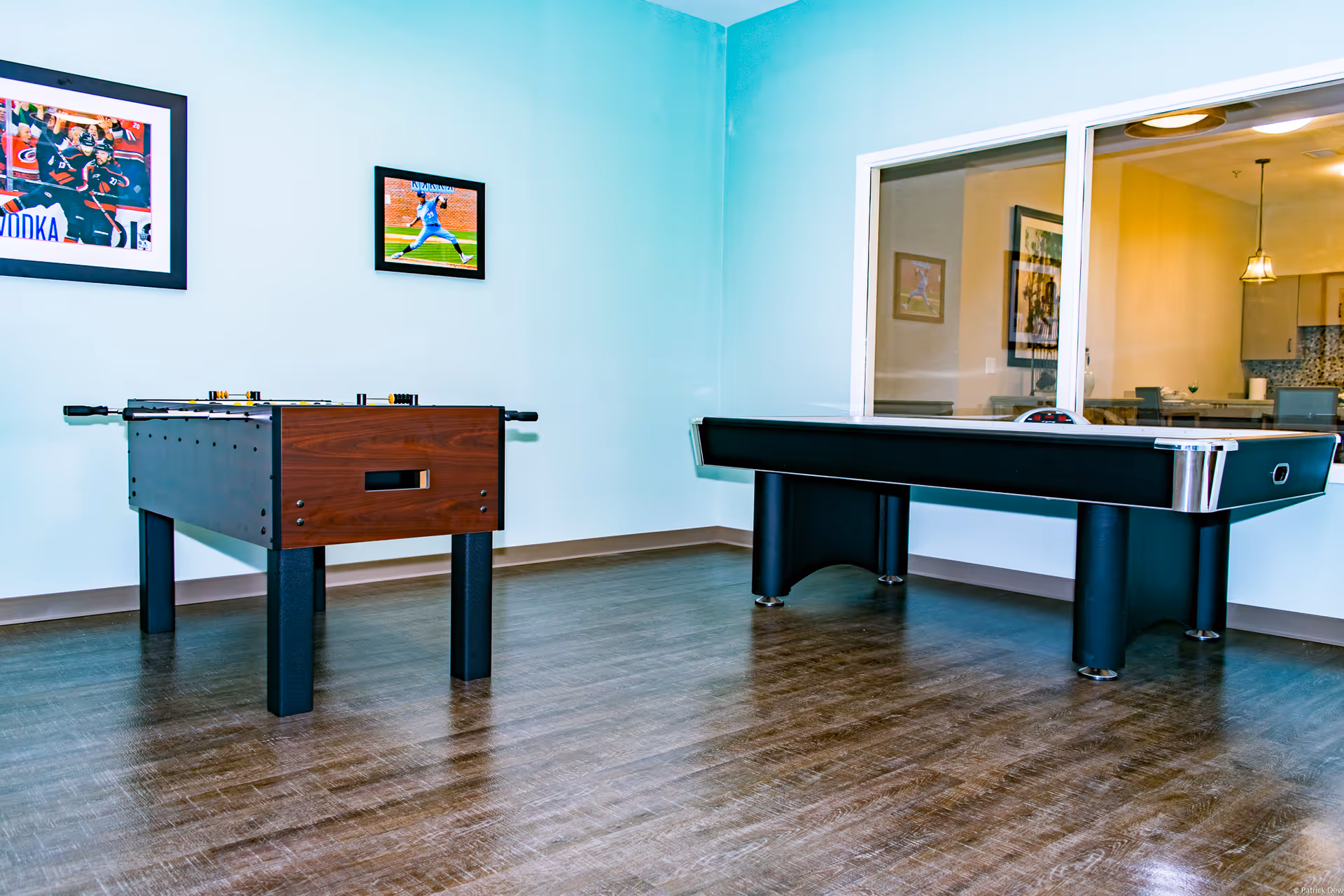A recreation room interior with a foosball table and a large game table against a pale blue wall and wood floor.